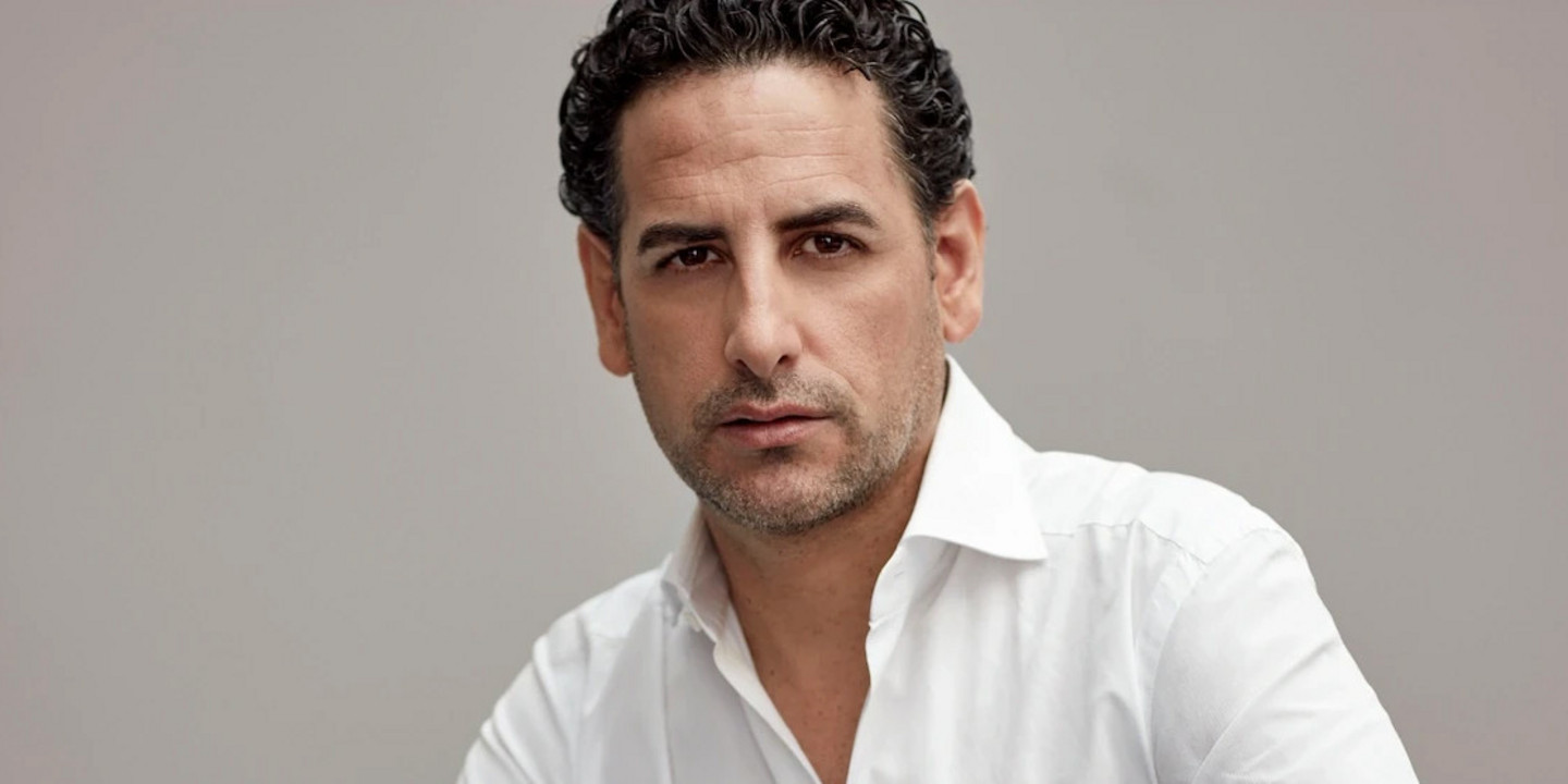 © Gregor Hohenberg Ein Porträt von Juan Diego Flórez, der in weißem Hemd und dunkler Hose posiert; er sitzt halb kniend vor neutralem Hintergrund und blickt ernst in die Kamera.