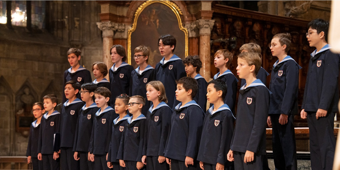 © Lukas Beck Die Wiener Sängerknaben in dunkelblauen Matrosenuniformen mit hellblauen Kragen singen in einer Kirche. Die Kinder stehen in mehreren Reihen vor einem kunstvoll gestalteten Altarraum mit goldenen und steinernen Elementen.