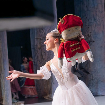 © www.coloss-art.com Eine Ballerina in weißem Tutu tanzt lächelnd auf der Bühne und hält eine große Stoffpuppe in roter Uniform, die an einen Nussknacker erinnert.