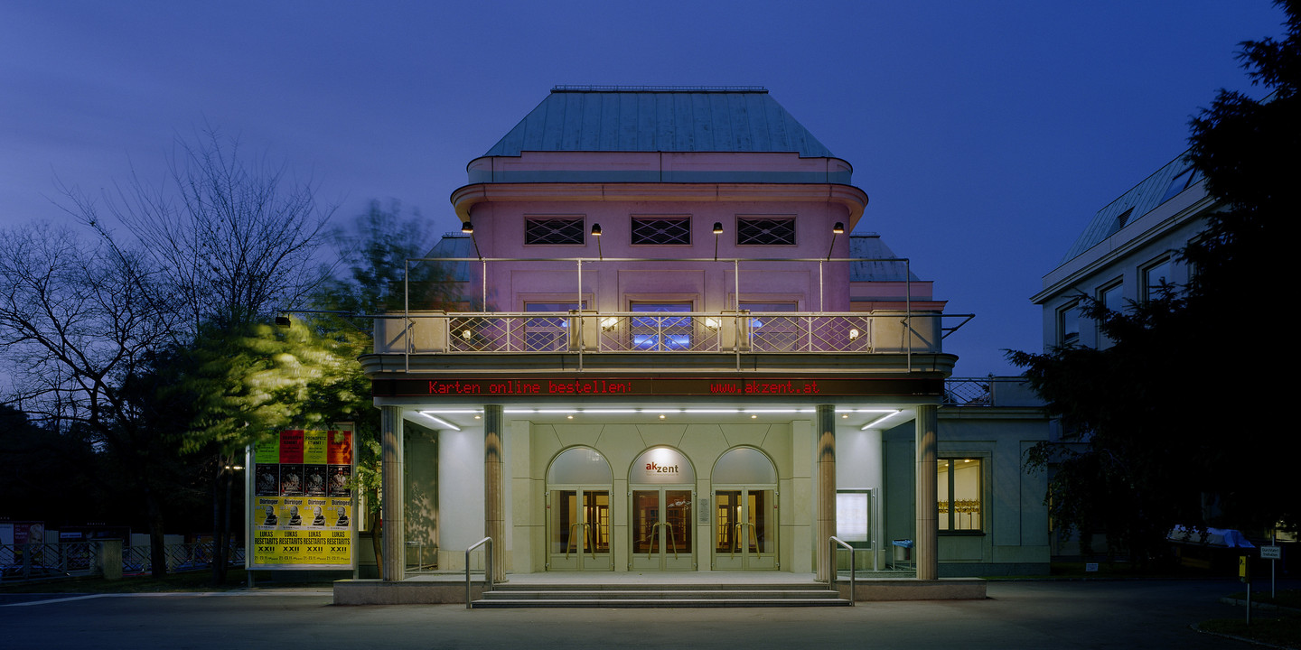 © Bruno Klomfar Abendliche Außenansicht des Theaters Akzent in Wien. Das Gebäude ist warm beleuchtet, die Fassade in zartem Rosa erstrahlt, und über dem Eingang leuchtet ein roter Schriftzug mit der Webadresse des Theaters.