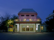 © Bruno Klomfar Abendliche Außenansicht des Theaters Akzent in Wien. Das Gebäude ist warm beleuchtet, die Fassade in zartem Rosa erstrahlt, und über dem Eingang leuchtet ein roter Schriftzug mit der Webadresse des Theaters.