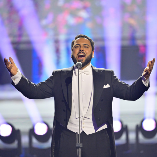 © ORF Ein Opernsänger in festlichem Frack singt leidenschaftlich auf einer hell erleuchteten Bühne, umgeben von bunten Lichteffekten und Scheinwerfern.