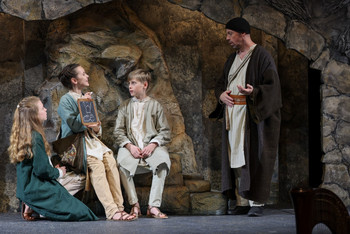 Drei Kinder in Gewändern zeigen eine Zeichnung auf einer Schiefertafel, während ein Mann in braunem Mantel mit ihnen spricht, Szene beim Salzburger Adventsingen.