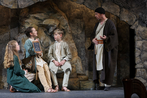 Drei Kinder in Gewändern zeigen eine Zeichnung auf einer Schiefertafel, während ein Mann in braunem Mantel mit ihnen spricht, Szene beim Salzburger Adventsingen.