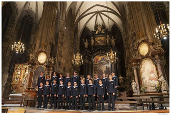 © Lukas Beck Die Wiener Sängerknaben in dunkelblauen Uniformen stehen aufgereiht im Altarraum einer prächtigen Kirche mit hohen Gewölben, vergoldeten Altären und leuchtenden Kronleuchtern.