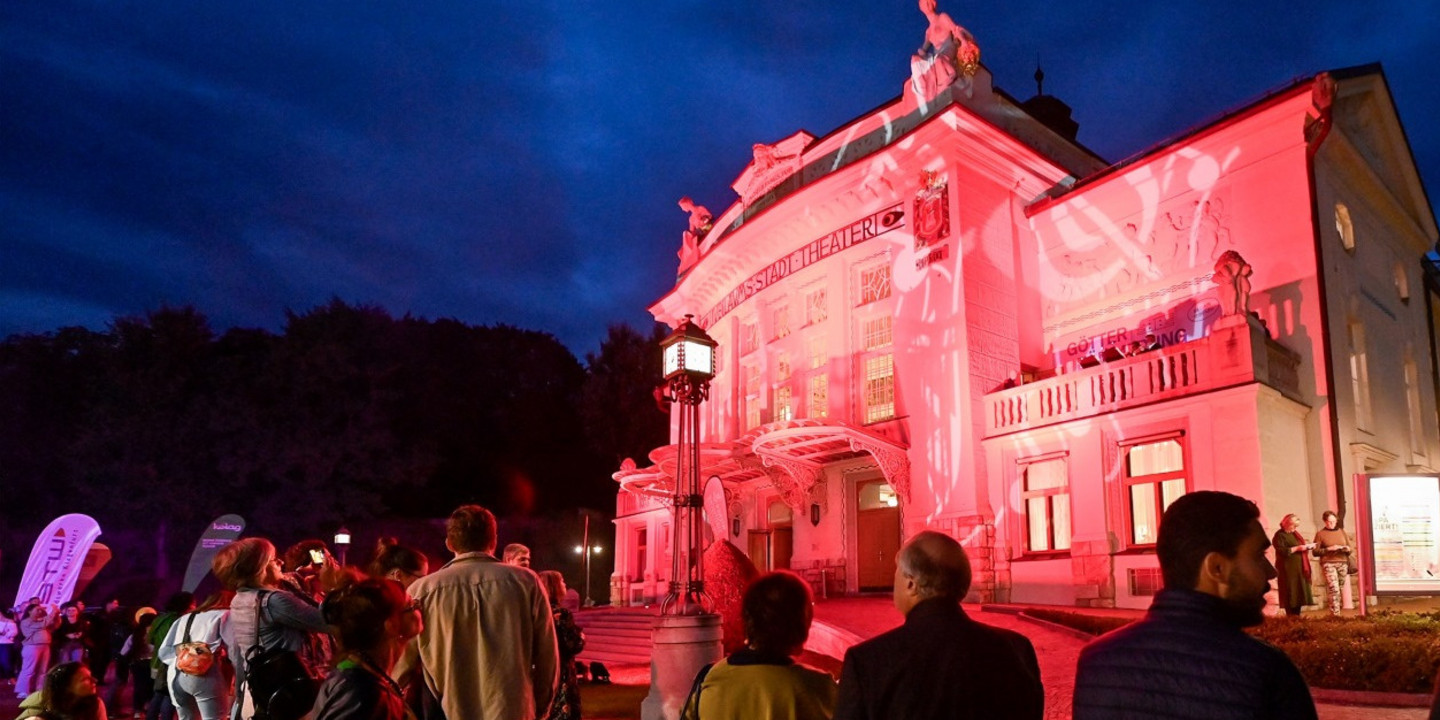 Menschen versammeln sich am Abend vor einem hellrot beleuchteten Theatergebäude. Das Gebäude ist kunstvoll verziert, und die Fassade wird durch farbiges Licht eindrucksvoll in Szene gesetzt. Der Himmel ist tiefblau, und die Besucher stehen auf dem gepflasterten Vorplatz und beobachten das Geschehen.