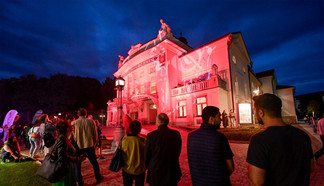 © Helge Bauer Menschen versammeln sich am Abend vor einem hellrot beleuchteten Theatergebäude. Das Gebäude ist kunstvoll verziert, und die Fassade wird durch farbiges Licht eindrucksvoll in Szene gesetzt. Der Himmel ist tiefblau, und die Besucher stehen auf dem gepflasterten Vorplatz und beobachten das Geschehen.