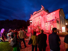 Menschen versammeln sich am Abend vor einem hellrot beleuchteten Theatergebäude. Das Gebäude ist kunstvoll verziert, und die Fassade wird durch farbiges Licht eindrucksvoll in Szene gesetzt. Der Himmel ist tiefblau, und die Besucher stehen auf dem gepflasterten Vorplatz und beobachten das Geschehen.