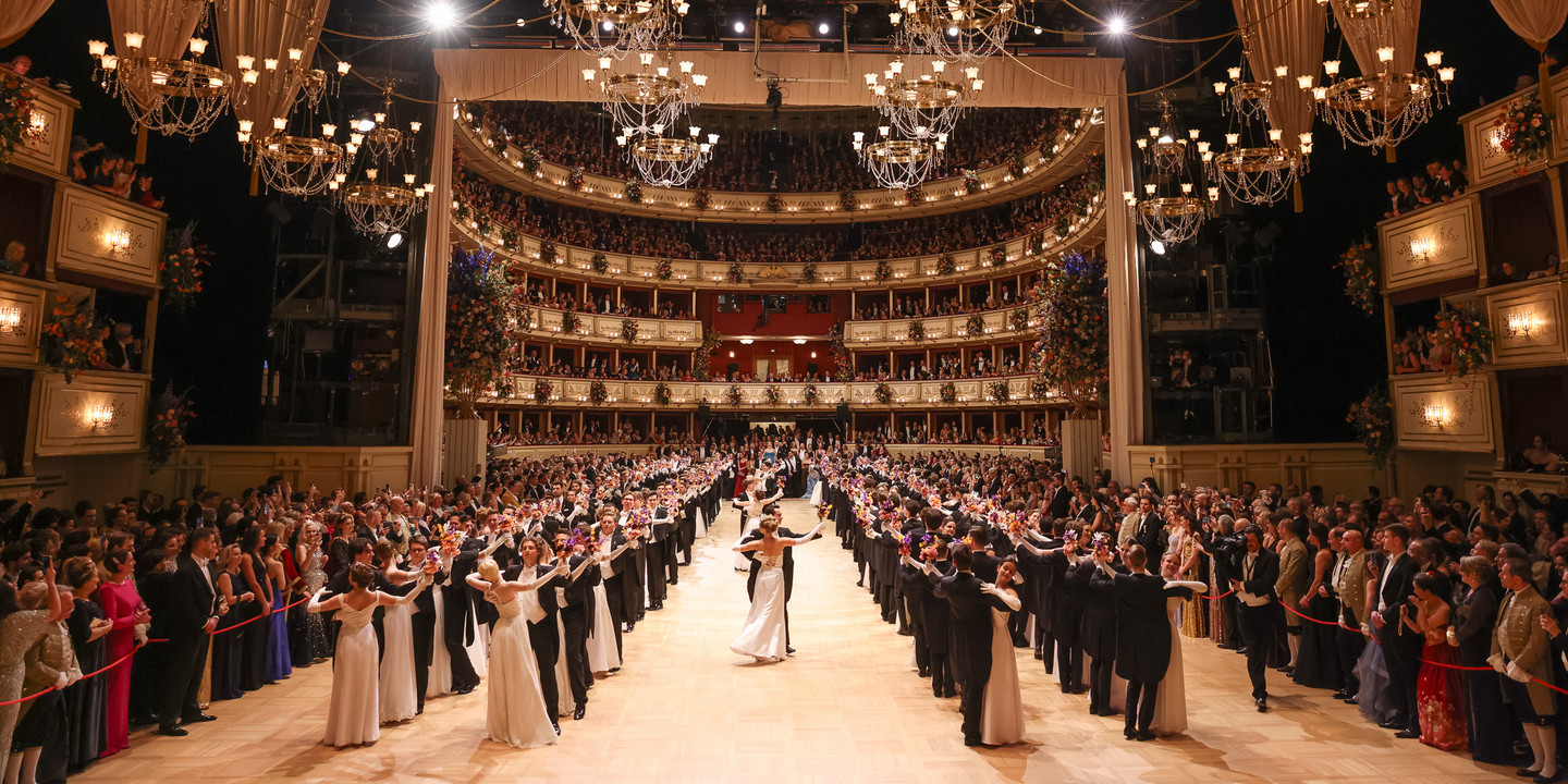 Festlich beleuchteter Opern- oder Ballsaal mit mehreren Kronleuchtern und voll besetzten Rängen. Auf dem Parkett tanzen zahlreiche Paare in Abendkleidern und Anzügen in einer symmetrischen Formation, umgeben von Publikum.
