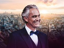 Ein Mann im Smoking mit Fliege lächelt vor einer Stadtansicht bei Sonnenuntergang.