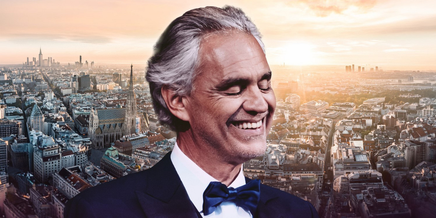 Ein Mann im Smoking mit Fliege lächelt vor einer Stadtansicht bei Sonnenuntergang.