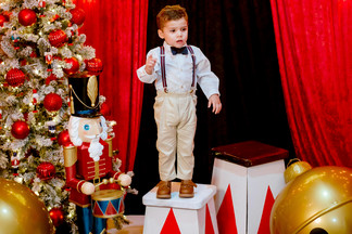 © Pexels, Luis Zambrano Ein kleiner Junge steht festlich gekleidet mit Hosenträgern und Fliege auf einer dekorativen Bühne neben einem Weihnachtsbaum und einem Nussknacker, umgeben von roten Vorhängen und goldenen Kugeln.