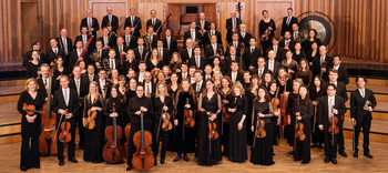 © Hans Leitner Das ORF Radio-Symphonieorchester Wien versammelt sich für ein offizielles Gruppenfoto auf einer Bühne mit Holzverkleidung; die Musiker:innen tragen elegante Konzertkleidung und halten ihre Instrumente.