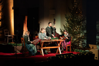 © www.salzburgeradvent.at Volksmusiker in Tracht sitzen an einem Tisch auf der Bühne vor großen, rot-blau beleuchteten Orgelpfeifen; Konzertszene im Saal.