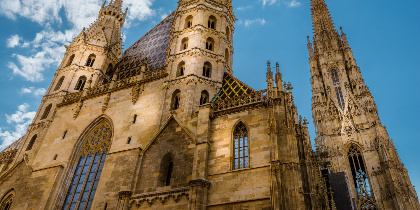 Der Stephansdom in Wien mit seinen gotischen Türmen und dem bunt gefliesten Dach vor blauem Himmel mit ein paar Wolken.

