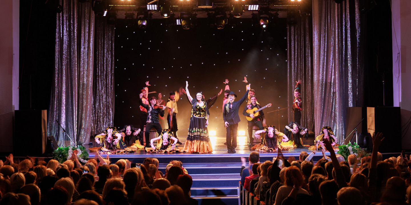 Bühnenaufführung mit farbenfroher Tanz- und Musikshow: Künstlerinnen und Künstler in bunten Kostümen tanzen und musizieren vor funkelndem Sternen-Hintergrund, während das Publikum begeistert applaudiert.