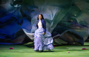 Eine Schauspielerin steht auf einer Bühne vor einem dramatisch beleuchteten, blauen Hintergrund aus weichen Stoffbahnen. Sie trägt eine weiße Bluse, eine blaue Jacke und einen weit ausgestellten Rock in hellblauen Tönen. Einzelne Blumen liegen auf dem grünen Bühnenboden verstreut.
