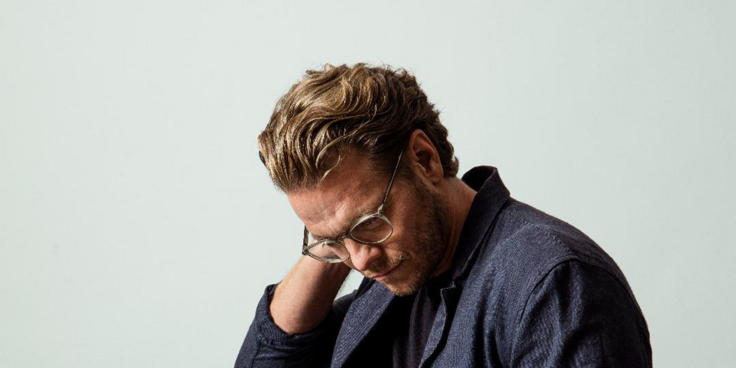 © Andrea Peller Ein Mann mit hellbraunen Haaren und Brille steht nachdenklich vor einem hellgrauen Hintergrund. Er trägt ein dunkelblaues Sakko über einem schwarzen T-Shirt und Jeans, hat eine Hand in der Hosentasche und blickt nach unten.