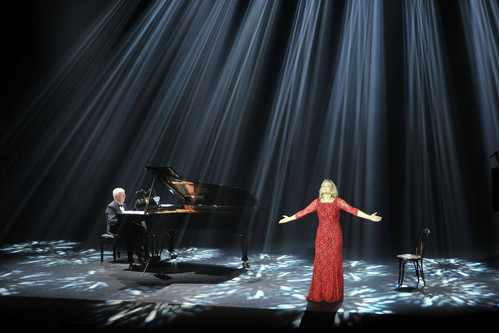 © Kristaps Kalns Sängerin im roten Kleid mit ausgebreiteten Armen auf einer beleuchteten Bühne, begleitet von einem Pianisten am Flügel.