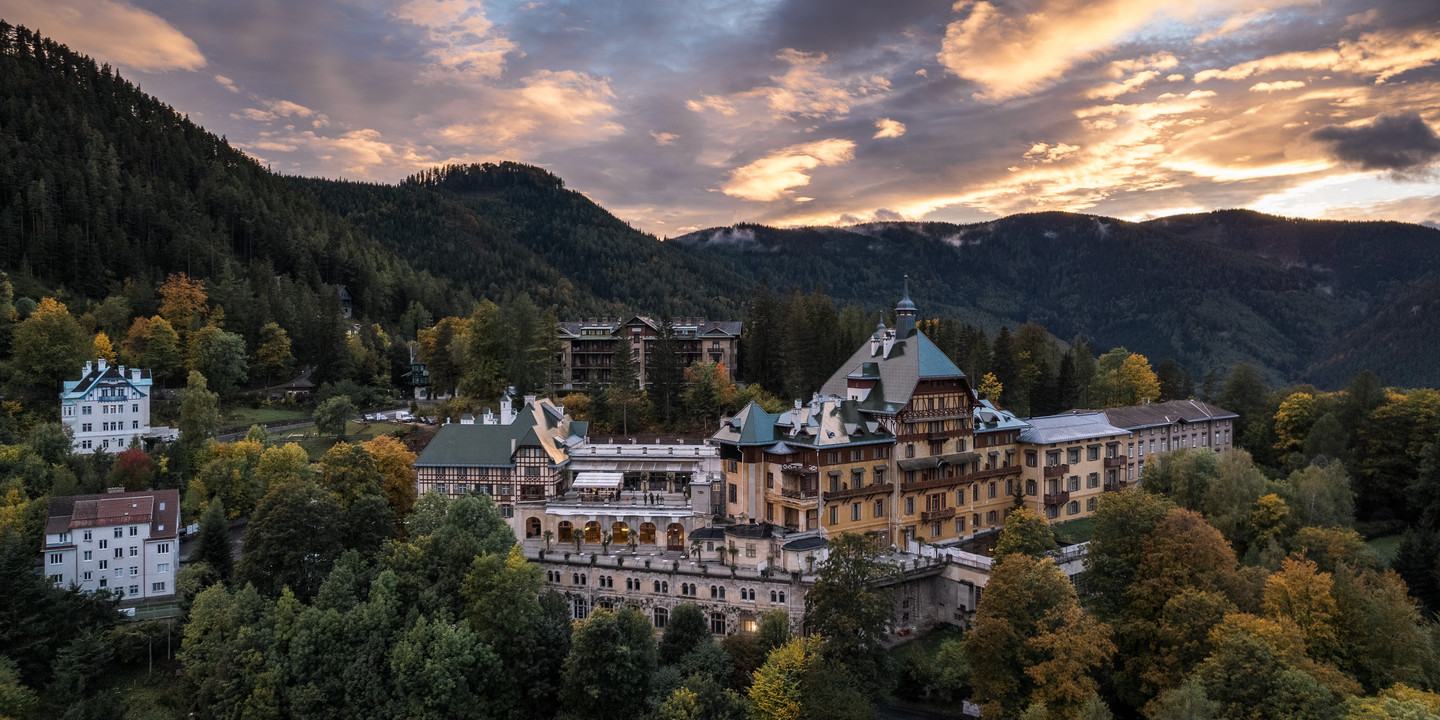 © Ian Ehm Historisches Grandhotel inmitten eines herbstlich gefärbten Waldes am Semmering, aufgenommen bei Sonnenuntergang mit dramatisch gefärbtem Himmel im Hintergrund.