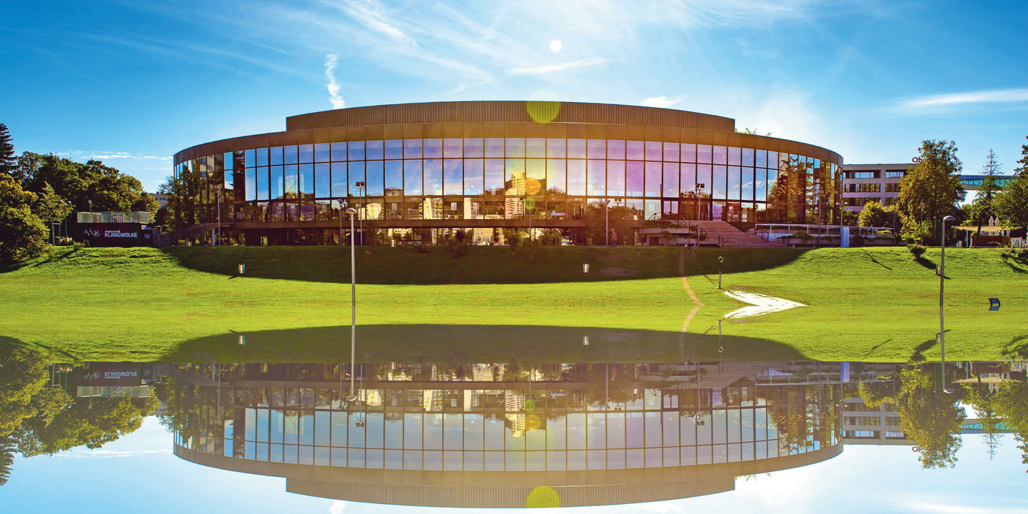 © Rita Newman Modernes Konzerthaus mit Glasfassade, davor grüne Wiese und Wasserreflexion.