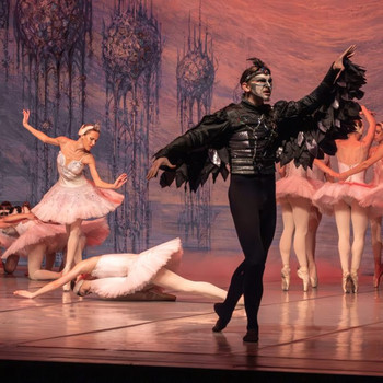 © www.coloss-art.com Eine Szene aus einem Ballett: Ein Tänzer in dunklem Kostüm mit Flügeln steht im Vordergrund, während Tänzerinnen in rosa Tutus elegant im Hintergrund posieren.