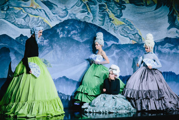 © Apollonia T. Bitzan Vier Schauspielerinnen in barocken Kostümen mit hohen weißen Perücken und Fächern auf einer Theaterbühne, vor einer gemalten Berglandschaft als Bühnenbild.