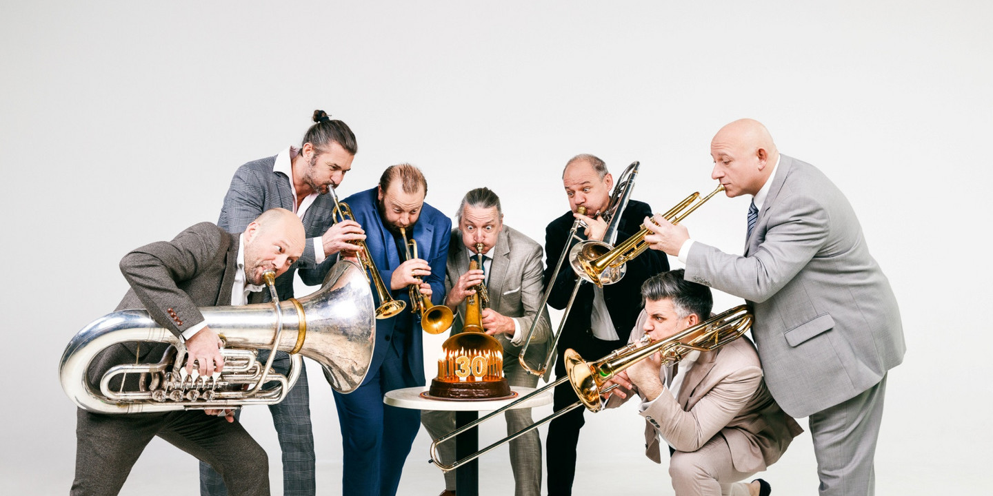 © Maria Frodl Sieben Männer in Anzügen stehen in einem Halbkreis um einen Tisch mit einer Geburtstagstorte und spielen ihre Blasinstrumente in Richtung der Torte. Jeder Musiker hält ein anderes Blechblasinstrument, die Szene wirkt humorvoll und lebendig, vor einem hellen Hintergrund.