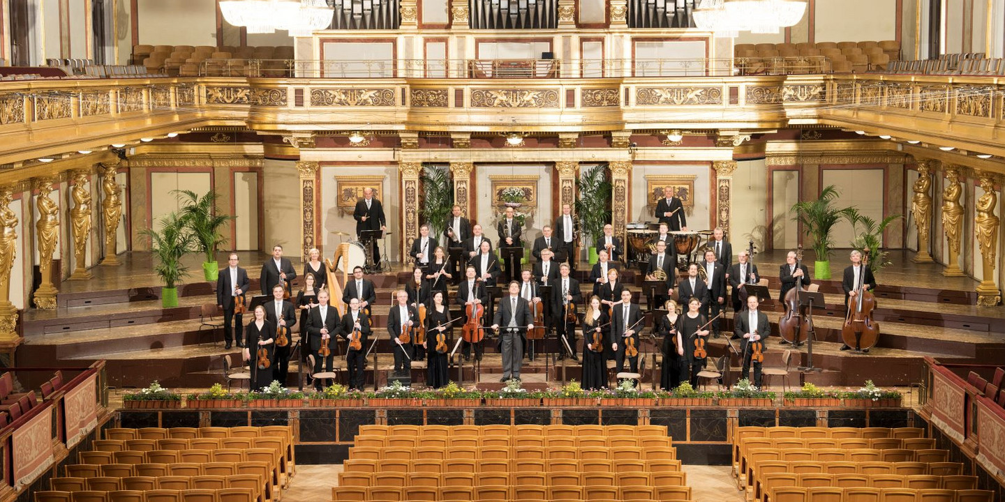 Ein klassisches Orchester steht auf der Bühne des Wiener Musikvereins, umgeben von goldverzierten Säulen, großen Kronleuchtern und der beeindruckenden Orgel im Hintergrund. Die Musikerinnen und Musiker halten ihre Instrumente und blicken in Richtung Publikum.
