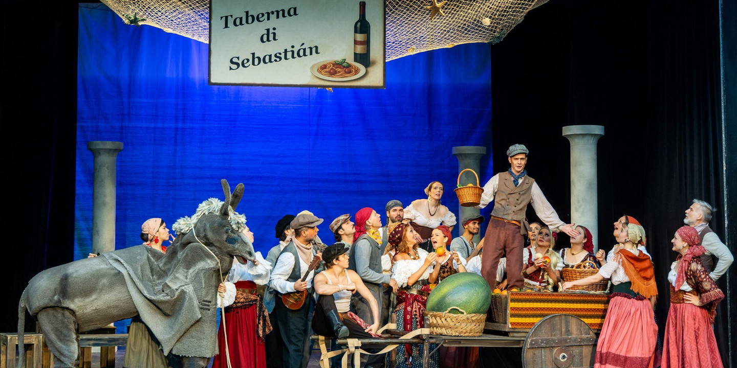 © Christian Husar Bühnenbild einer italienisch inspirierten Theaterszene mit vielen Darsteller:innen in bunten, historischen Kostümen vor einem blauen Hintergrund.