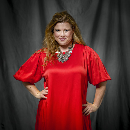 © Stephan Doleschal Porträt einer Frau in einem auffälligen roten Kleid mit großen Puffärmeln, die selbstbewusst lächelt und die Hände in die Hüften stemmt. Der Hintergrund ist dunkel und schlicht.