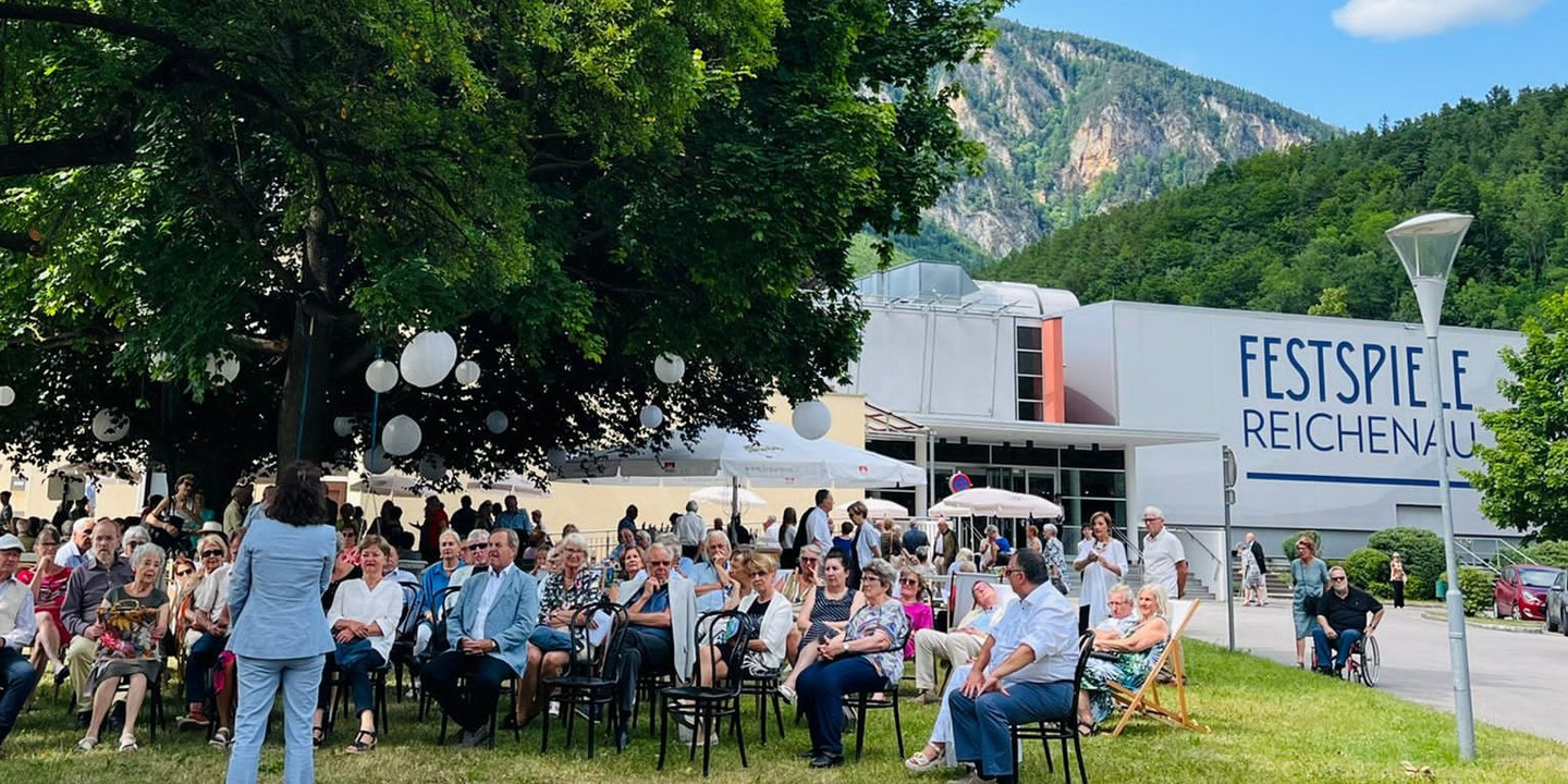 Publikum sitzt unter einem schattenspendenden Baum auf einer Wiese vor dem Festspielhaus Reichenau, im Hintergrund Berge und blauer Himmel mit vereinzelten Wolken, sommerliche Festivalatmosphäre.
