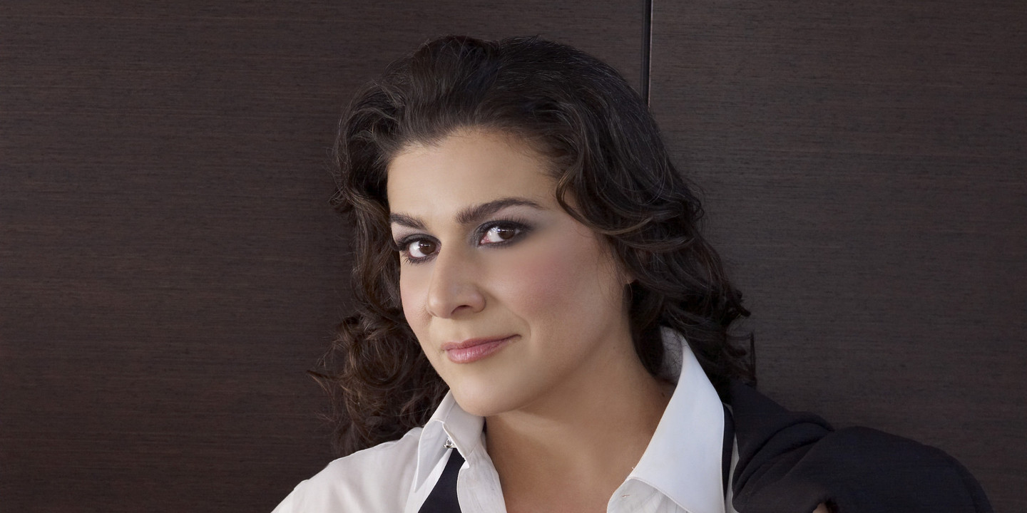 Cecilia Bartoli im weißen Hemd mit offenem Smokinghemd und schwarzer Fliege, die Jacke lässig über die Schulter gelegt, vor dunklem Hintergrund.