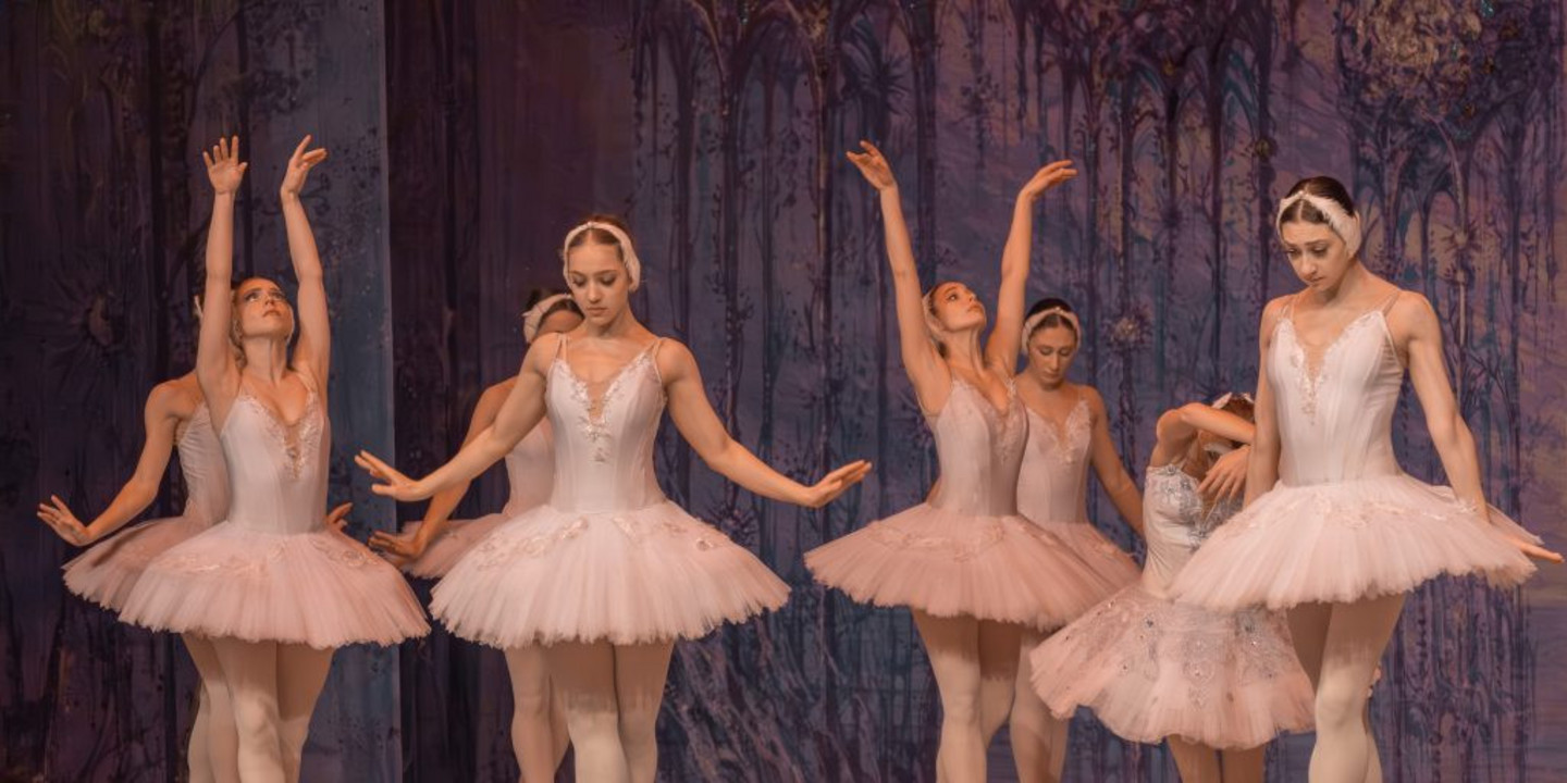 © www.coloss-art.com Eine Gruppe von Ballerinen in weißen Tutus tanzt elegant auf Spitze. Sie stehen in anmutigen Posen vor einem violett beleuchteten Bühnenhintergrund.
