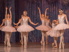 © www.coloss-art.com Eine Gruppe von Ballerinen in weißen Tutus tanzt elegant auf Spitze. Sie stehen in anmutigen Posen vor einem violett beleuchteten Bühnenhintergrund.