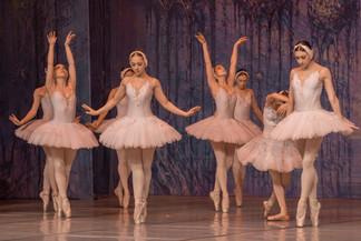 Eine Gruppe von Ballerinen in weißen Tutus tanzt elegant auf Spitze. Sie stehen in anmutigen Posen vor einem violett beleuchteten Bühnenhintergrund.