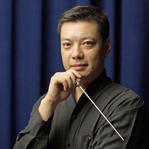 © Wu Promotion Ein Dirigent hält einen Taktstock und blickt selbstbewusst in die Kamera vor einem dunklen Bühnenvorhang.