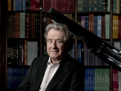 © Marco Borggreve Porträt des Pianisten Rudolf Buchbinder vor einem schwarzen Flügel, im Hintergrund ein Bücherregal mit farbigen Buchrücken.