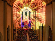 © Klaus Huber Stimmungsvolle Innenaufnahme einer Kirche mit festlicher Lichtprojektion, Weihnachtsbäumen und Chor auf der Bühne – die warme Beleuchtung schafft eine feierliche, besinnliche Atmosphäre.