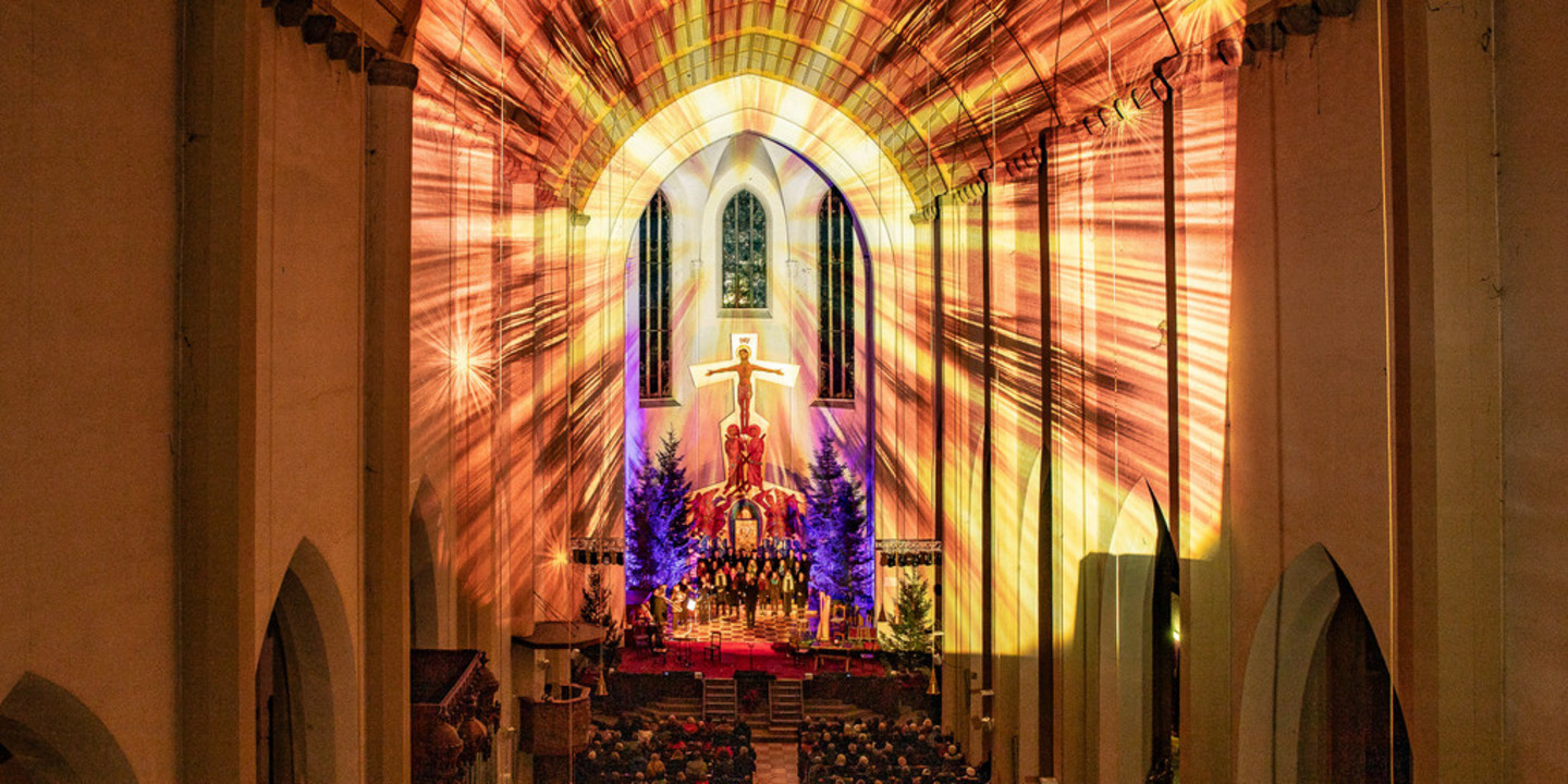 © Klaus Huber Stimmungsvolle Innenaufnahme einer Kirche mit festlicher Lichtprojektion, Weihnachtsbäumen und Chor auf der Bühne – die warme Beleuchtung schafft eine feierliche, besinnliche Atmosphäre.