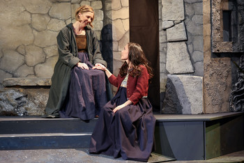 Zwei Darstellerinnen in historischen Kleidern sitzen auf einer steinernen Kulisse und halten sich die Hände, eine innige Szene beim Salzburger Adventsingen.