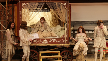 © Moritz Schell Eine Theaterszene in historischen Kostümen: Vier Darsteller stehen um eine reich verzierte Schlafkoje auf Rädern, in der eine Figur im Bett liegt, während die anderen mit überzeichneten Gesten agieren.