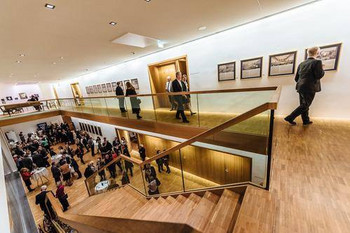 © Rudi Dellinger Innenansicht eines modernen Veranstaltungsgebäudes mit hellen Holzböden und Galerien. Besucher betrachten Kunstwerke an den Wänden und unterhalten sich im Foyer.