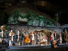 Junge Musiker:innen in traditionellen Gewändern musizieren mit Geigen, Kontrabass, Zither, Akkordeon und Gitarre vor einer Bühnenkulisse mit Weinreben und Bergen beim Salzburger Adventsingen.