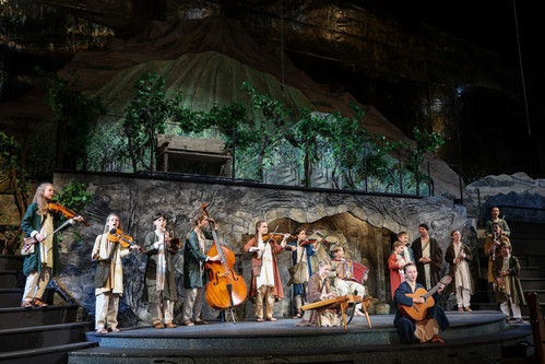 Junge Musiker:innen in traditionellen Gewändern musizieren mit Geigen, Kontrabass, Zither, Akkordeon und Gitarre vor einer Bühnenkulisse mit Weinreben und Bergen beim Salzburger Adventsingen.