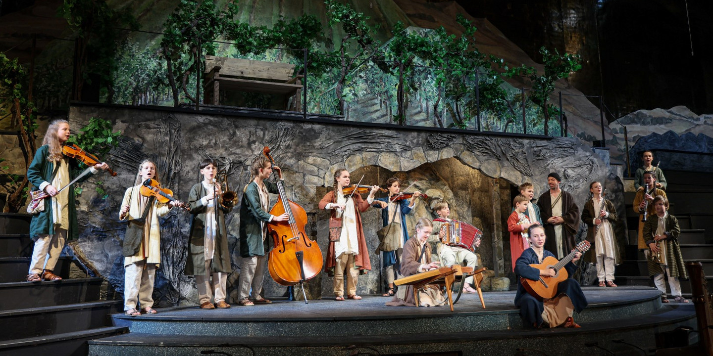 Junge Musiker:innen in traditionellen Gewändern musizieren mit Geigen, Kontrabass, Zither, Akkordeon und Gitarre vor einer Bühnenkulisse mit Weinreben und Bergen beim Salzburger Adventsingen.
