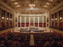 Großes Sinfoniekonzert in einem prunkvollen historischen Konzertsaal mit Orchester auf der Bühne und voll besetztem Publikum.