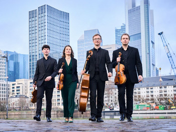 © Kaupo Kikkas Vier Musiker:innen des Eliot Quartetts in dunkler Konzertkleidung schreiten mit ihren Streichinstrumenten selbstbewusst über das Frankfurter Mainufer, im Hintergrund moderne Hochhäuser und Baustellenkräne der Skyline.