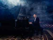 Ein Pianist sitzt in einem eleganten Konzertsaal an einem schwarzen Flügel und spielt konzentriert. Um ihn herum liegt leichter Bühnennebel, der die Atmosphäre geheimnisvoll wirken lässt. Der Saal ist leer, das Licht fällt weich auf die roten Sitzreihen und die kunstvoll gestalteten Wände im Hintergrund.