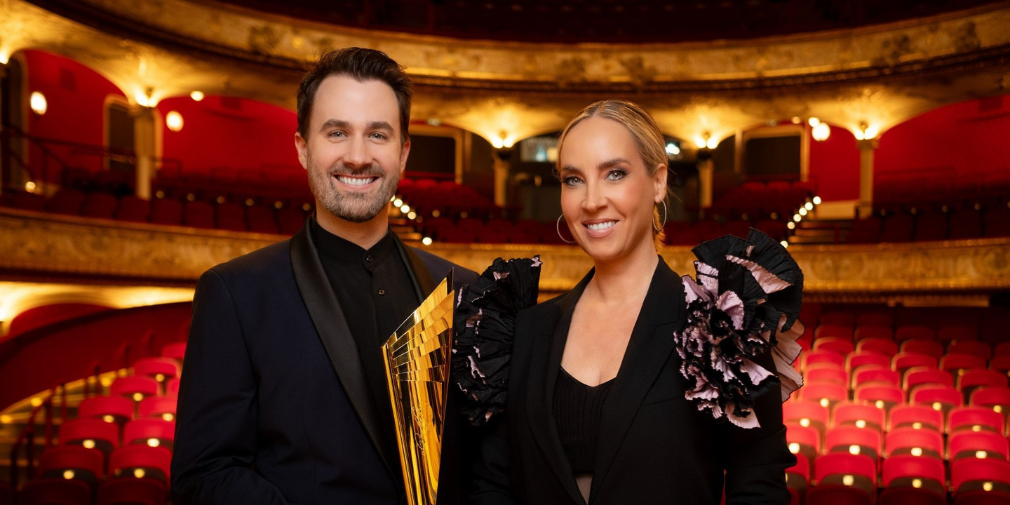Ein Mann und eine Frau in eleganter schwarzer Abendgarderobe stehen lächelnd in einem prachtvollen Theater mit rotem Samtinterieur. Der Mann hält eine goldene Trophäe in den Händen.